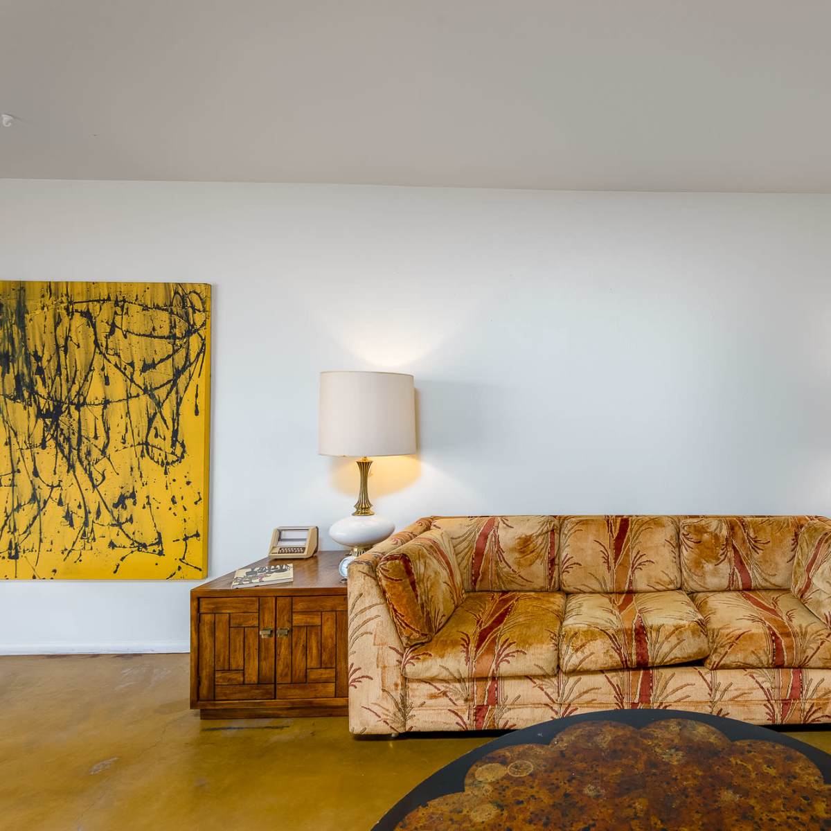 A living room featuring a patterned sofa, two table lamps, a wooden cabinet, and an abstract painting on the wall.