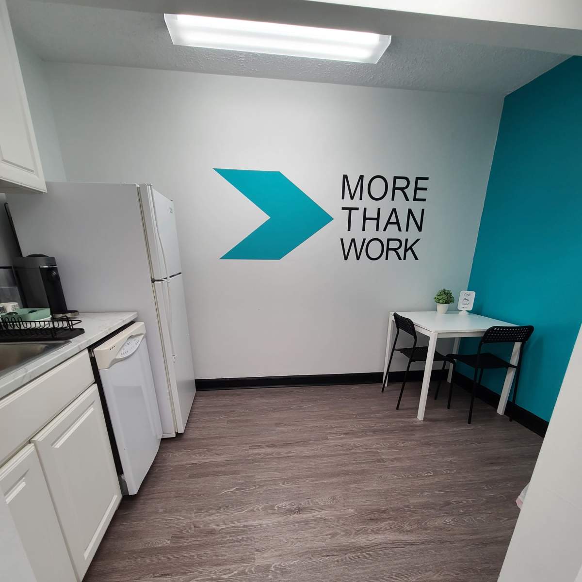 A kitchen area with white cabinets, a refrigerator, and a small table with two chairs, featuring a turquoise accent wall with the text "MORE THAN WORK."