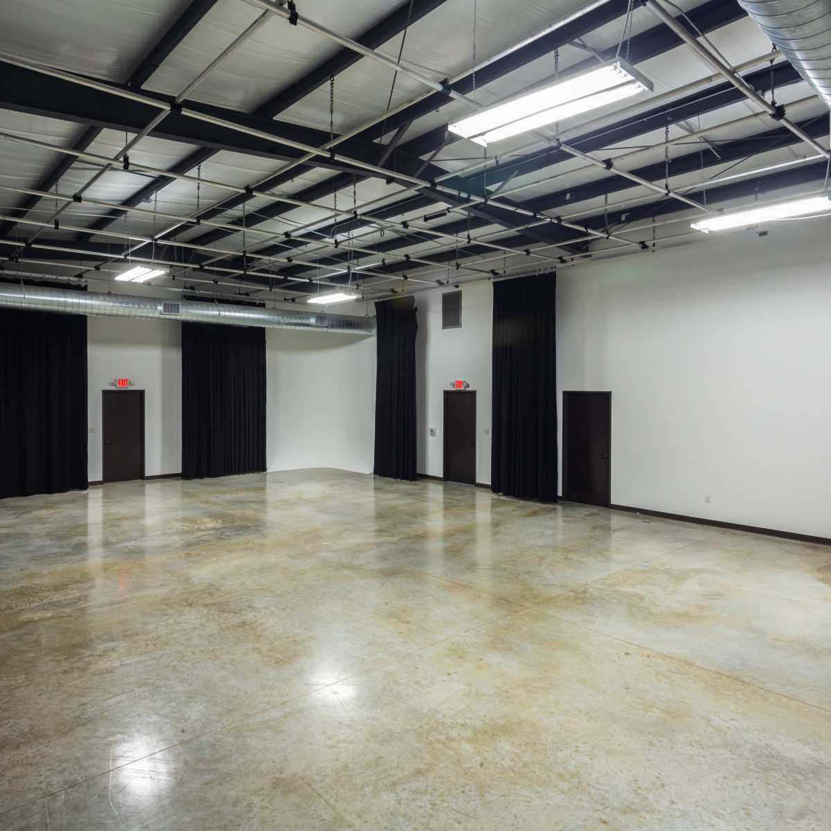 A large, empty room with concrete flooring, black curtains along the walls, and two doors on opposite sides.