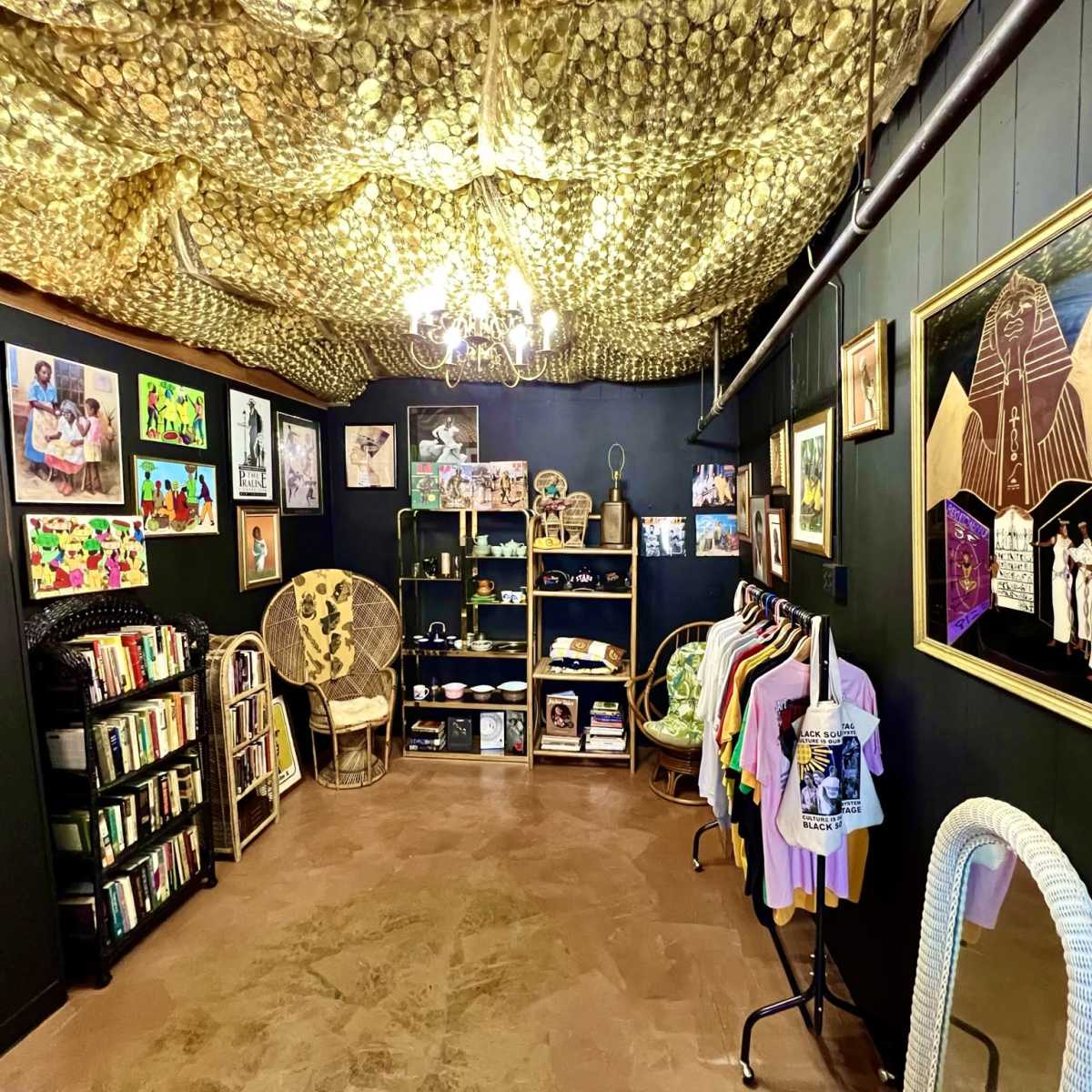 A cozy room with a ceiling draped in gold fabric, lined with framed artwork on the dark walls, shelves displaying various items, and clothing hanging on racks.