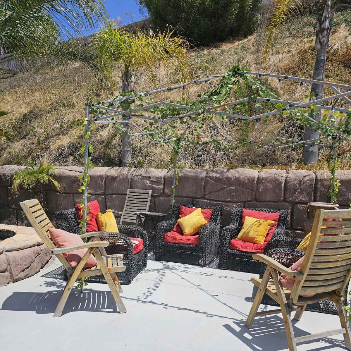 The scene features a modern outdoor seating area with two wooden chairs and a set of four cushioned chairs arranged under a vine-covered pergola.