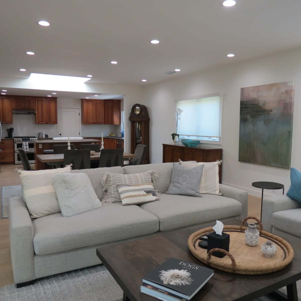 A modern living room features a light gray sectional sofa, a coffee table with decorative items, and an open layout leading to a kitchen area with wooden cabinets.