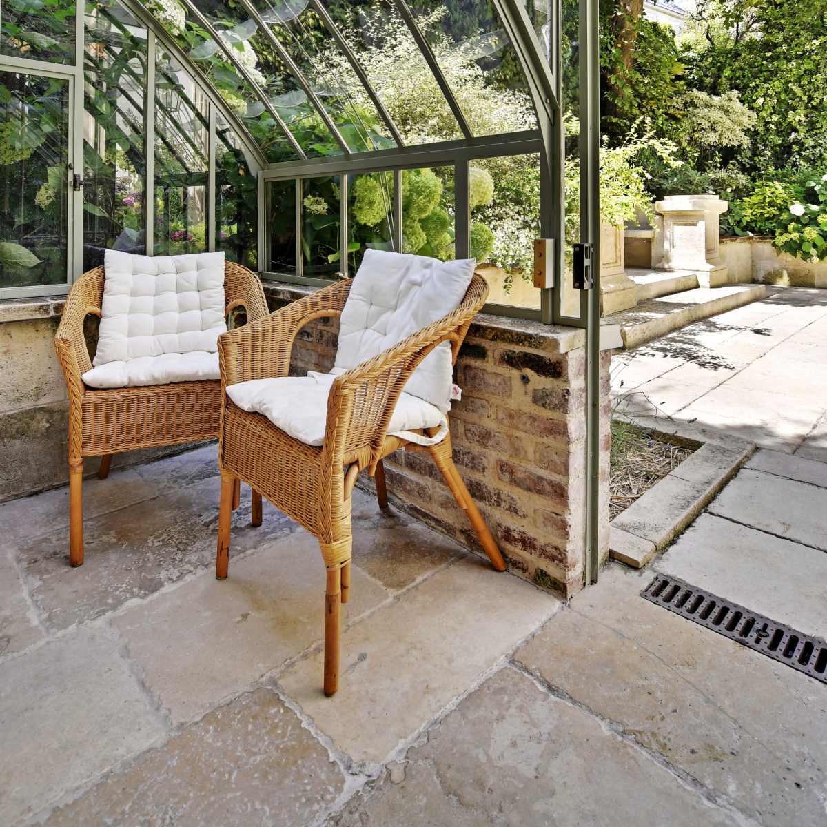 A conservatory features two wicker chairs with cushions, surrounded by lush greenery and stone flooring.
