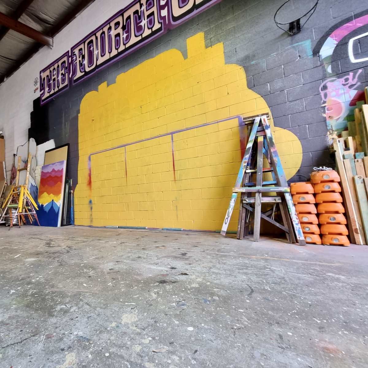 A spacious workshop features a bright yellow mural on the wall, with shelving units holding various supplies and tools stacked neatly to one side.