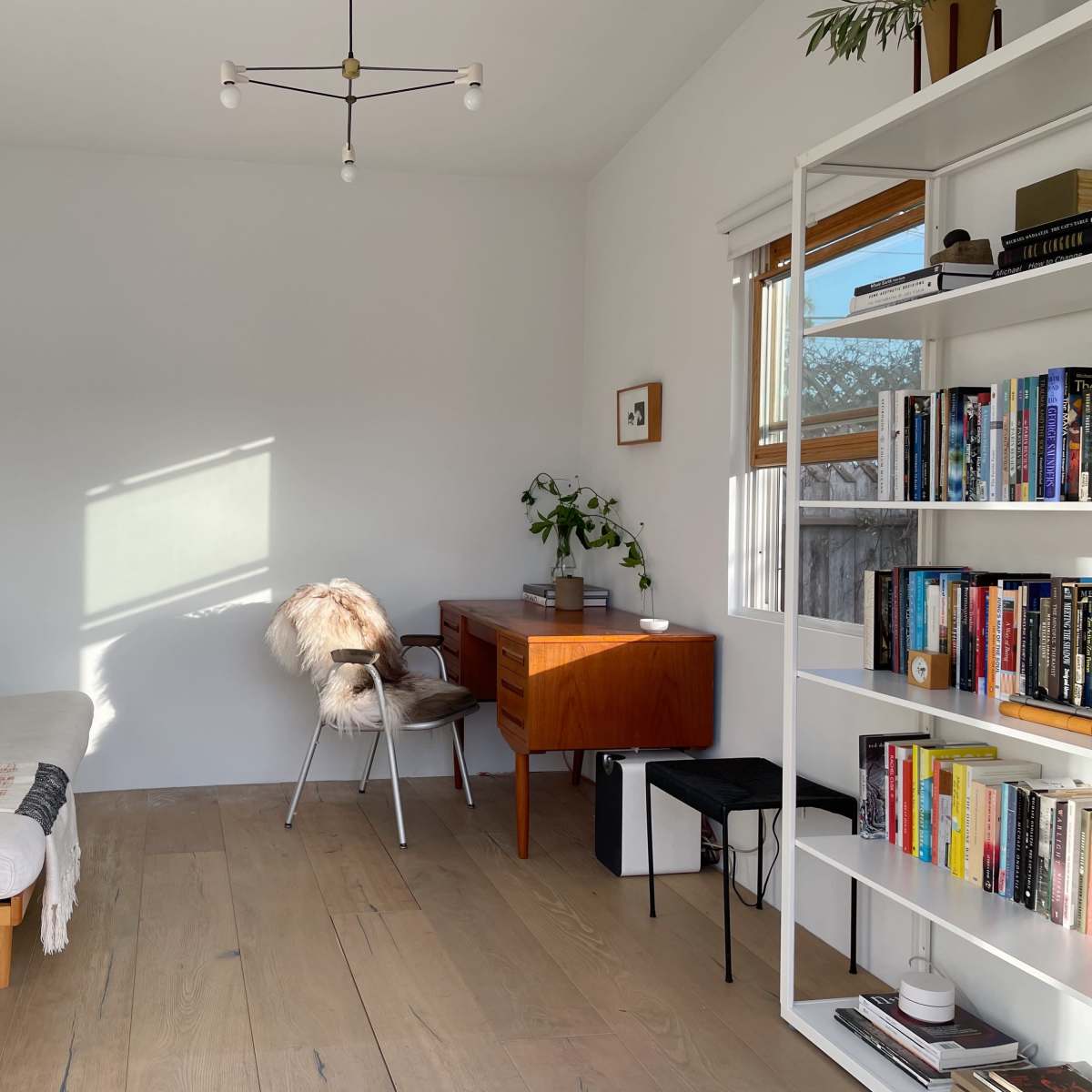 A bright, minimalist living room with a white sofa, a wooden desk, and a bookshelf filled with books.