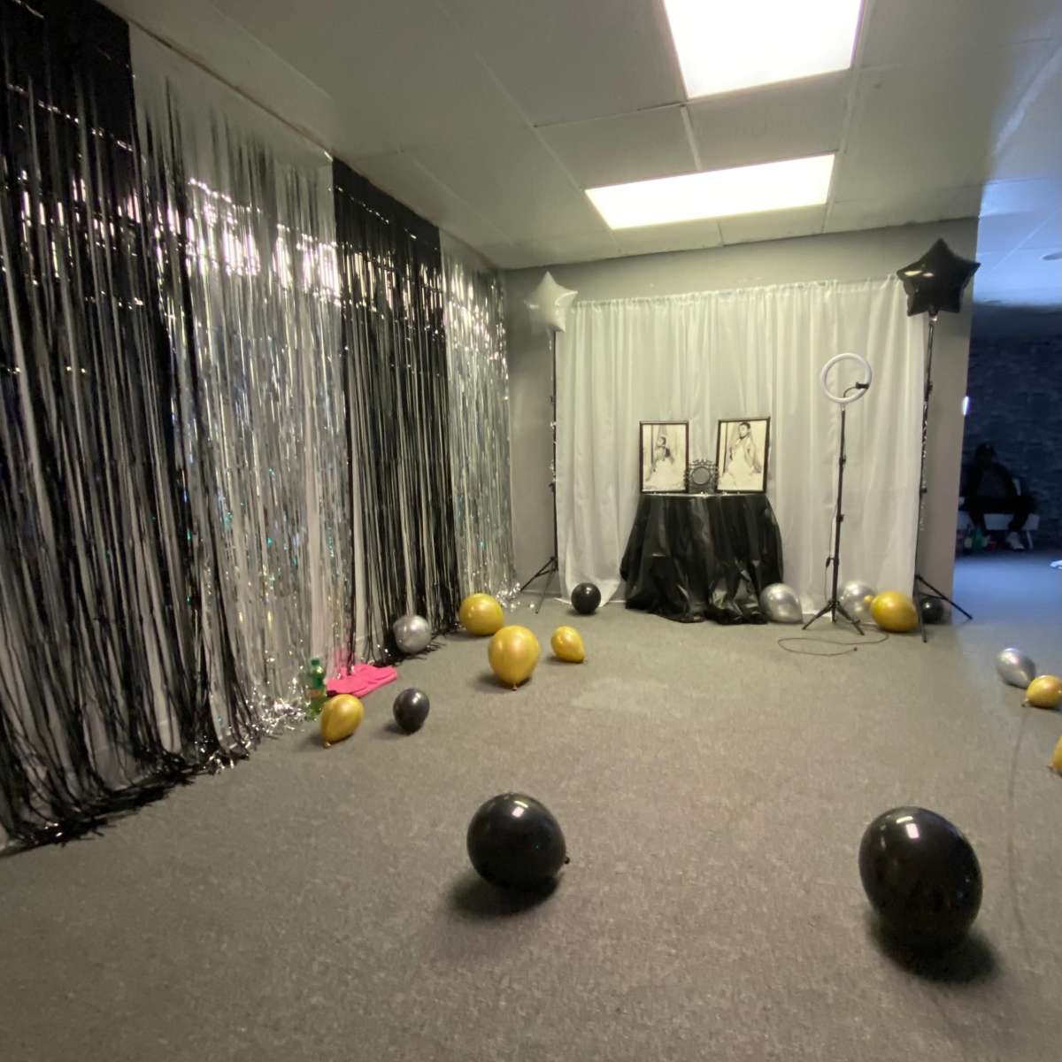 A party space decorated with black and silver fringe curtains, balloons in black and gold scattered on the floor, and a display featuring two framed pictures on a table draped in black cloth.