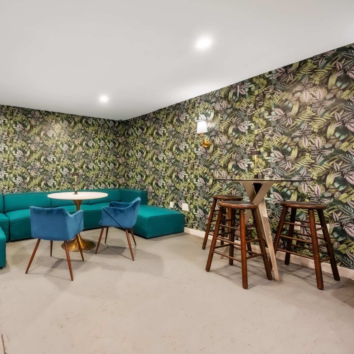 A modern lounge features a turquoise sectional sofa, a round table, and wooden stools against a vibrant, leafy wallpaper.