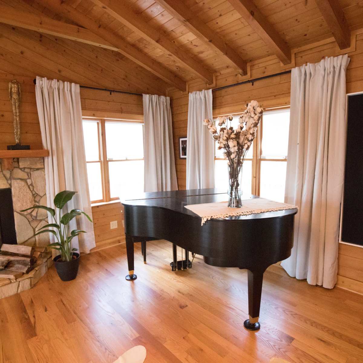 A grand piano sits in a wooden room beside a stone fireplace, with large windows framed by curtains and a decorative wall art nearby.