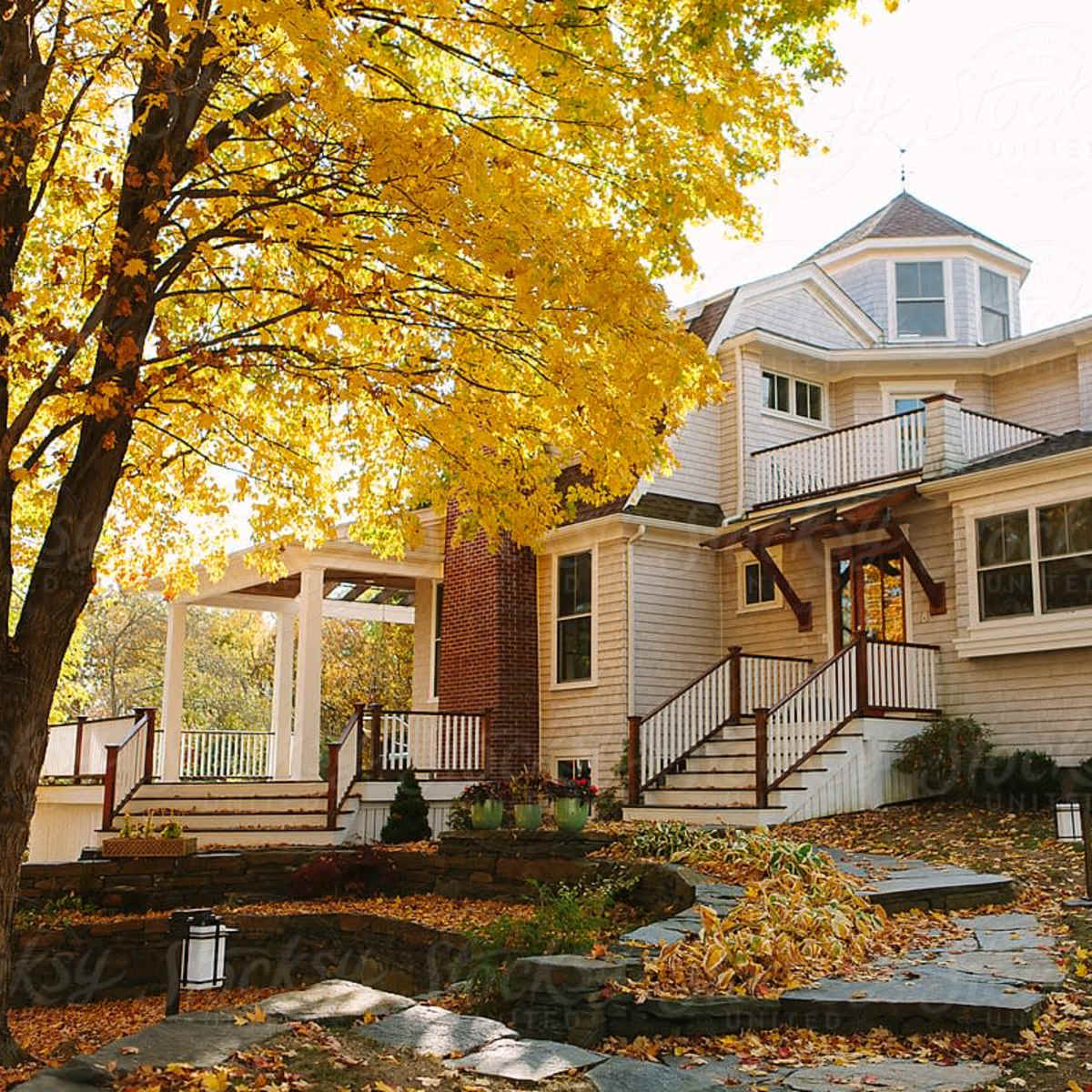 A two-story house with a light-colored facade and a porch is surrounded by trees displaying vibrant autumn foliage.