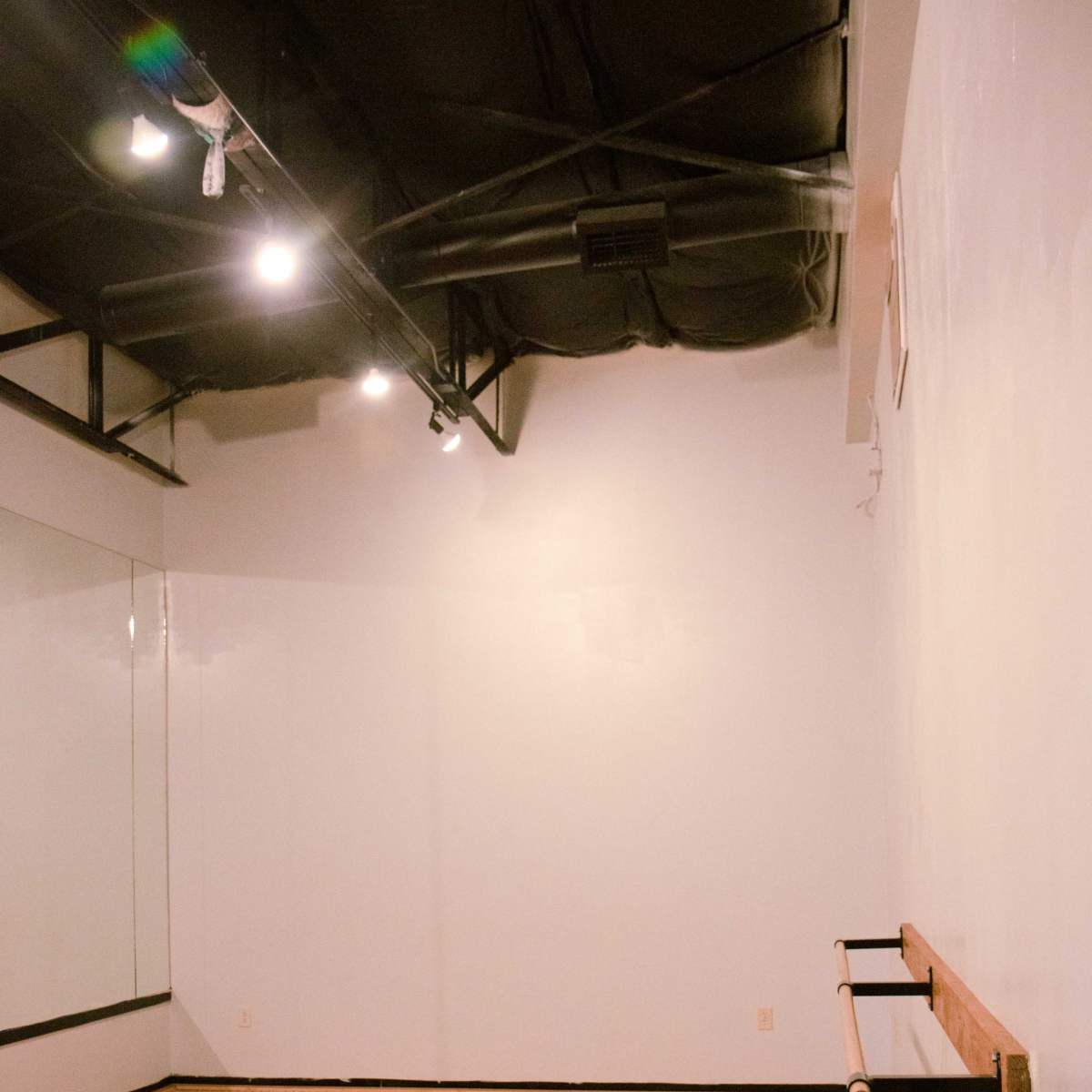 A dance studio with wooden flooring, a wall-mounted ballet barre, and a mirror partially visible on one wall.