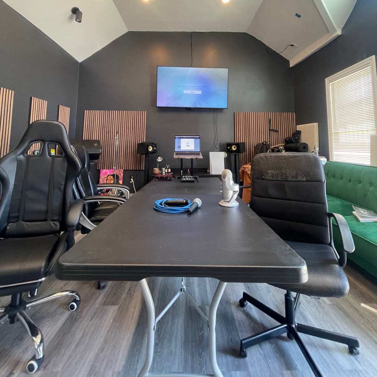 A recording studio with a large table, two black chairs, a computer setup, and acoustic panels on the walls.