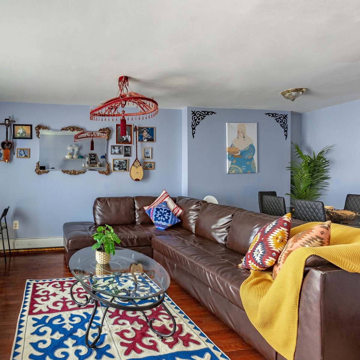 The living room features a brown leather sofa, a glass coffee table, colorful wall decor, and a large window with sheer curtains.