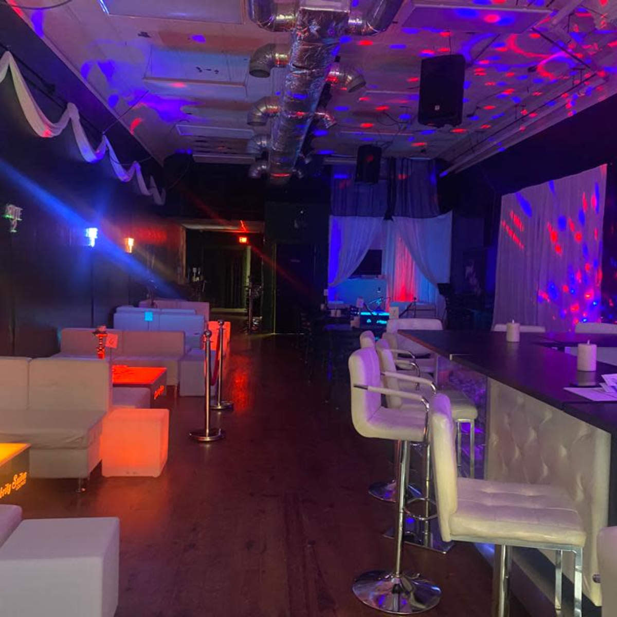 A dimly lit bar area with white lounge seating, illuminated by colorful stage lights and decorations.