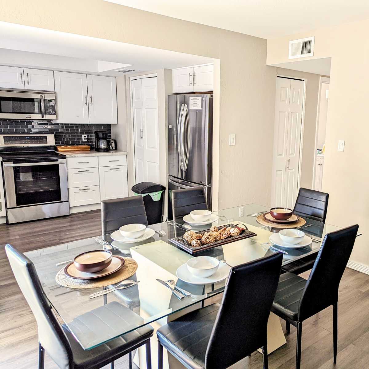 A modern dining room with a glass table set for six and a kitchen visible in the background.