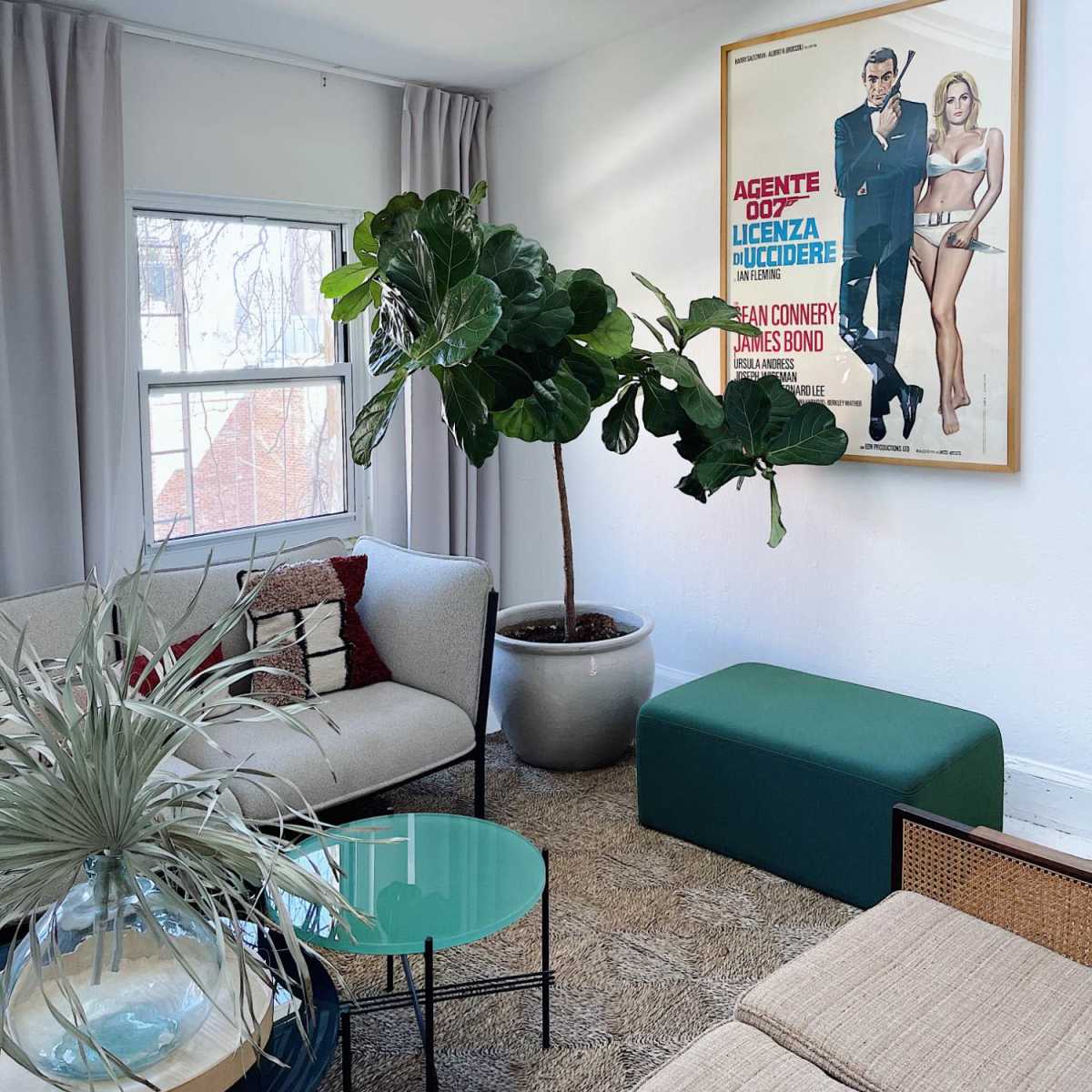 A living room features a framed movie poster, a large potted plant, a green bench, and modern furniture against a backdrop of natural light from a skylight.