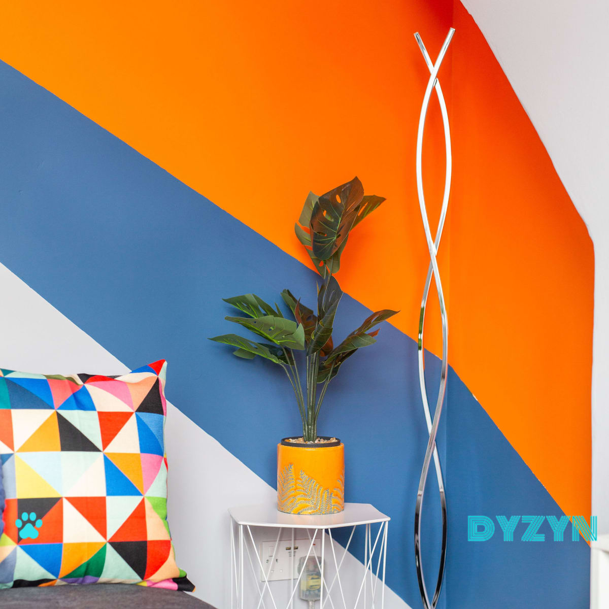 A corner of a room featuring an orange and blue accent wall, a geometric-patterned pillow on a gray sofa, a potted plant on a white side table, and a modern floor lamp.