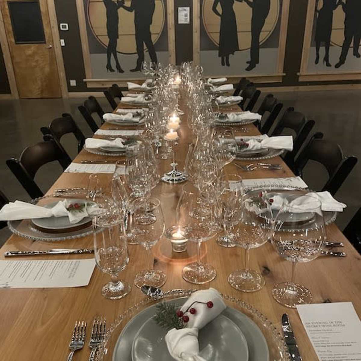 A long wooden dining table is elegantly set with fine china, glassware, and white napkins, surrounded by chairs in a room featuring silhouette artwork on the walls.