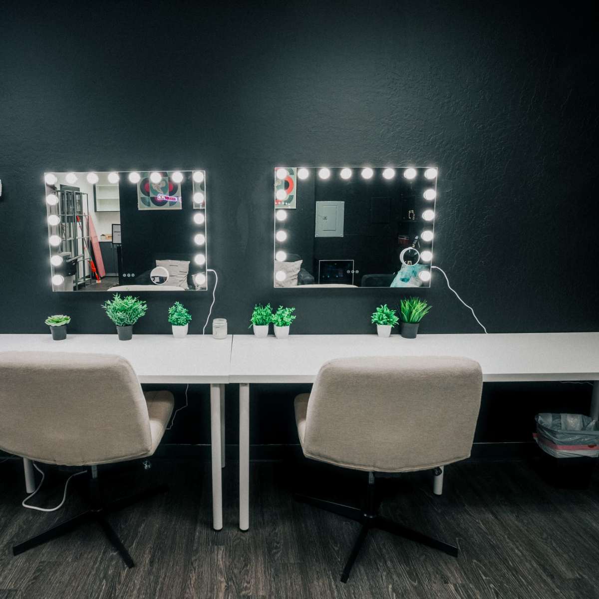 A modern workspace with two mirrored vanities, each framed by light bulbs, flanked by small potted plants, and a neon "ON AIR" sign on the wall.