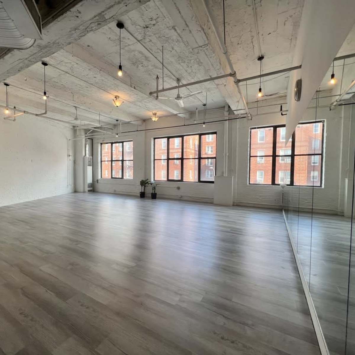 A spacious, minimalist room with large windows, wooden floors, and exposed ceilings, featuring a mirrored wall.