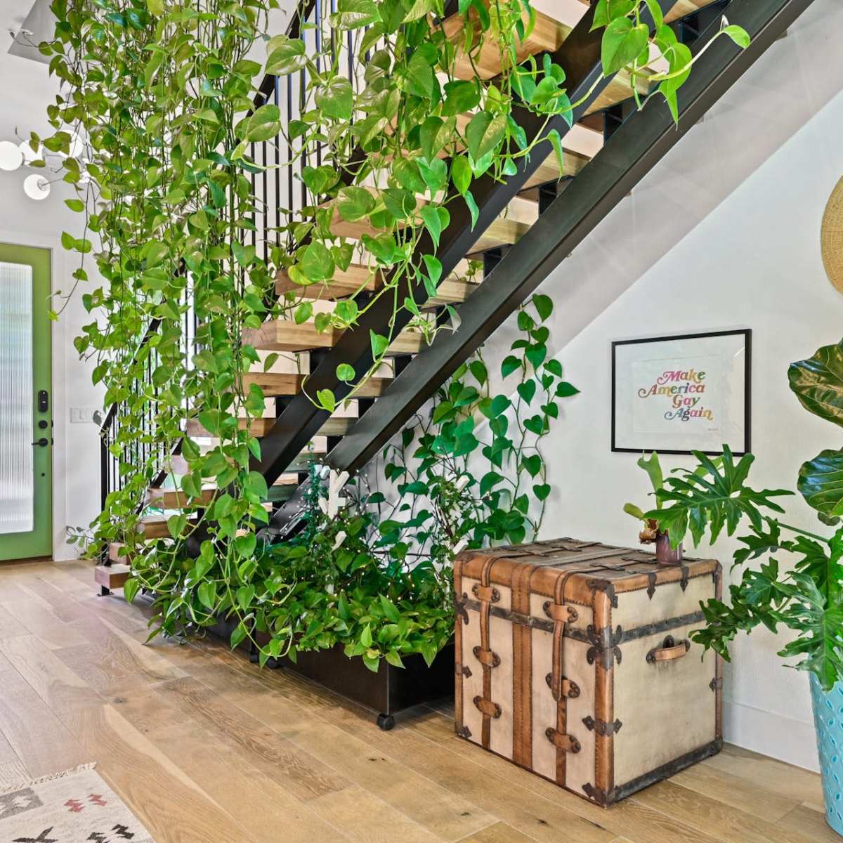 A staircase adorned with cascading green plants leads to a doorway, while a vintage trunk and a potted plant sit on the floor beside it.