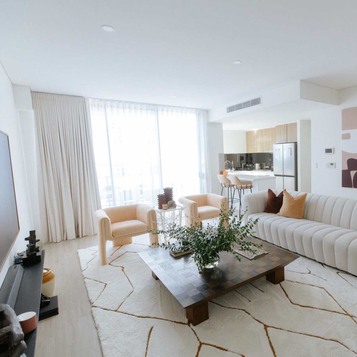 A modern living room features a beige sofa, two orange chairs, a wooden coffee table with a floral arrangement, and large windows with light-colored curtains.