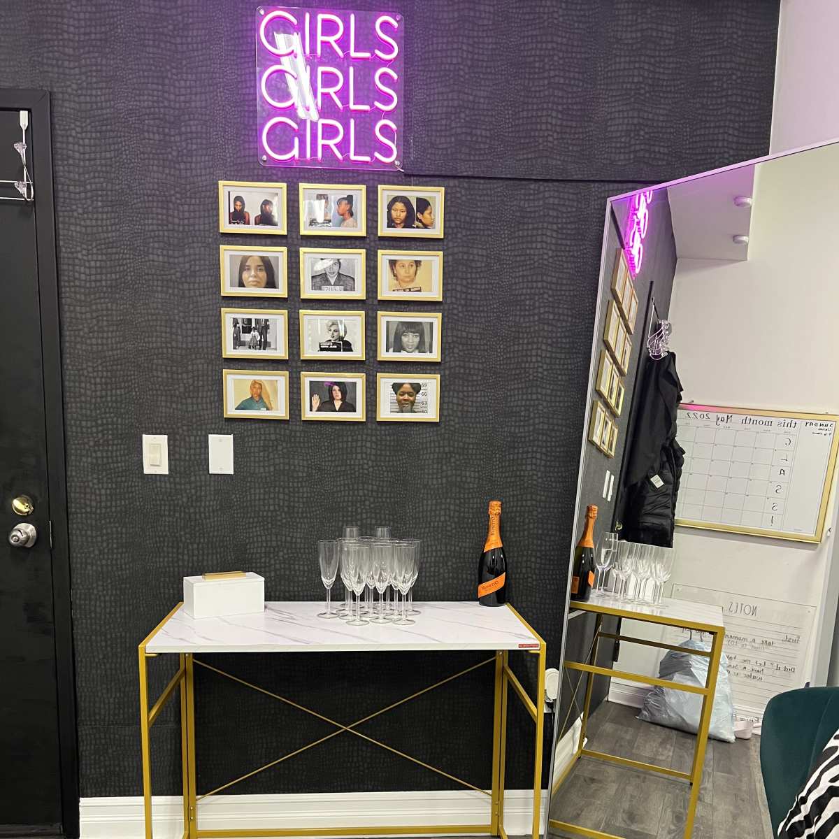 A small entryway features a marble-top table with champagne glasses and a bottle, alongside a mirror and framed portraits on a dark wall with a neon sign that reads "GIRLS."