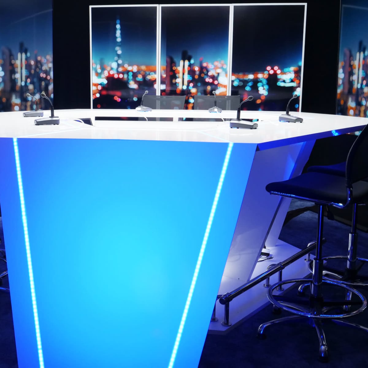 A modern broadcast studio with a brightly lit blue hexagonal table and several black barstools against a backdrop of a city skyline at night.