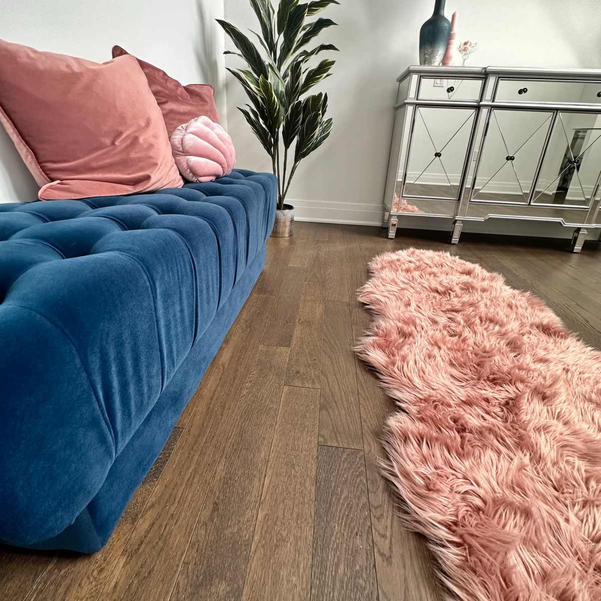 A blue tufted sofa is positioned next to a decorative plant and a mirrored console table on a hardwood floor, accompanied by a pink shag rug.