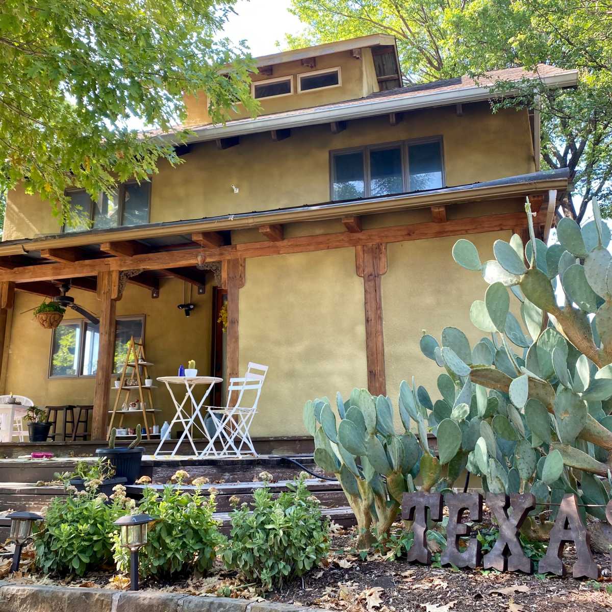 A two-story house with a greenish-yellow exterior is surrounded by trees and features a small outdoor seating area with a table and chairs, alongside a cactus garden and a wooden sign spelling "TEXAS."