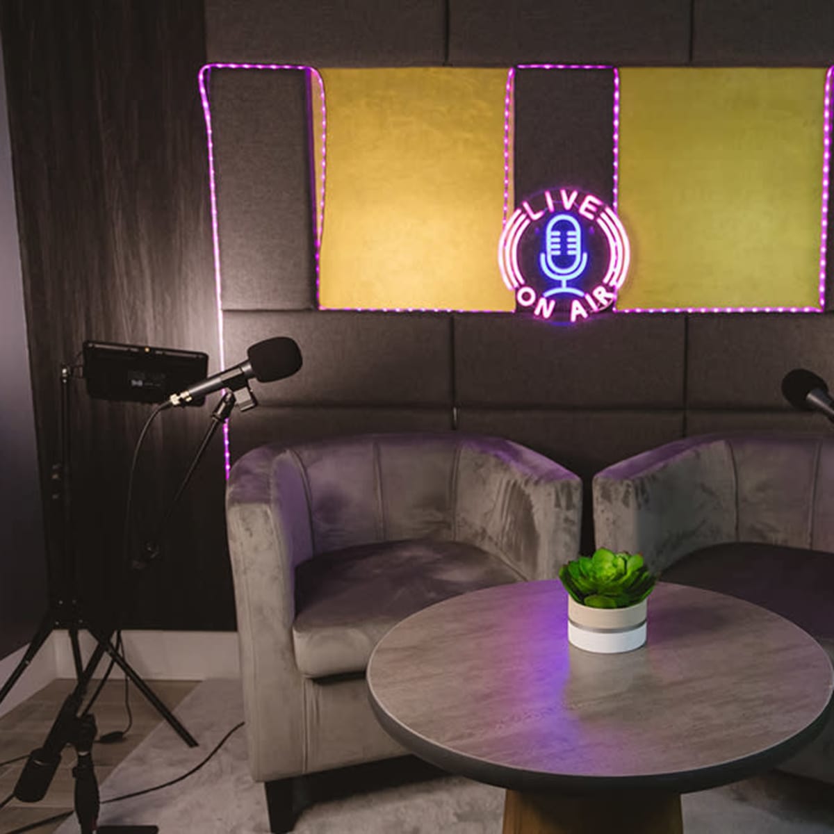 A cozy podcast studio features two chairs, a small table, and a "LIVE ON AIR" neon sign illuminated against a backdrop of padded wall panels.