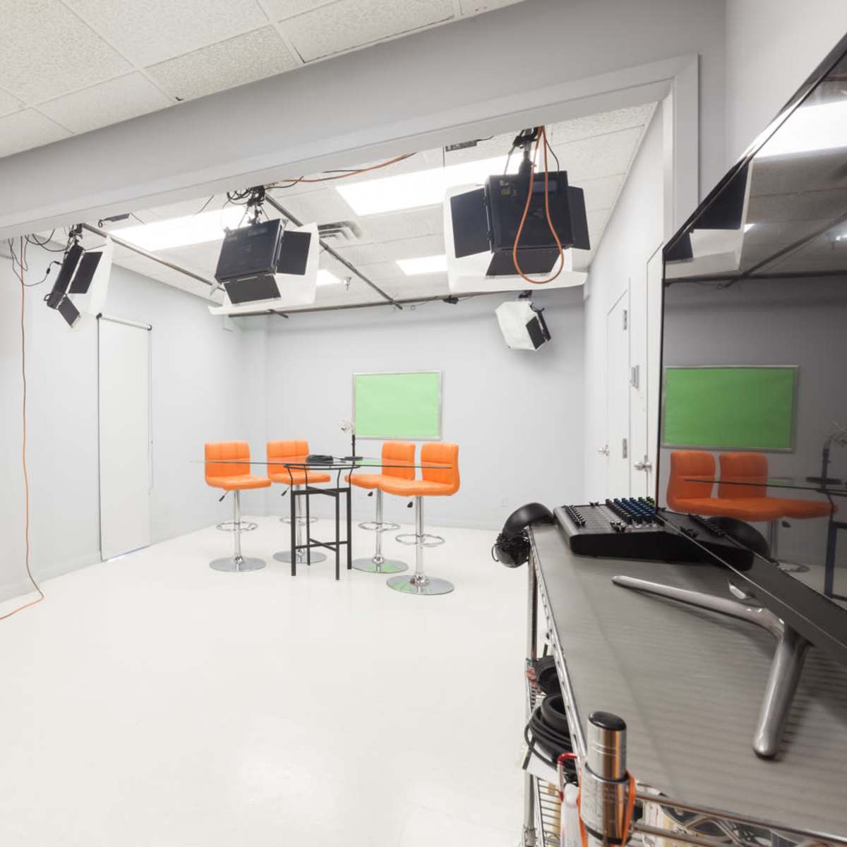 A modern studio space features orange bar stools around a black table, with professional lighting equipment and green screen areas.