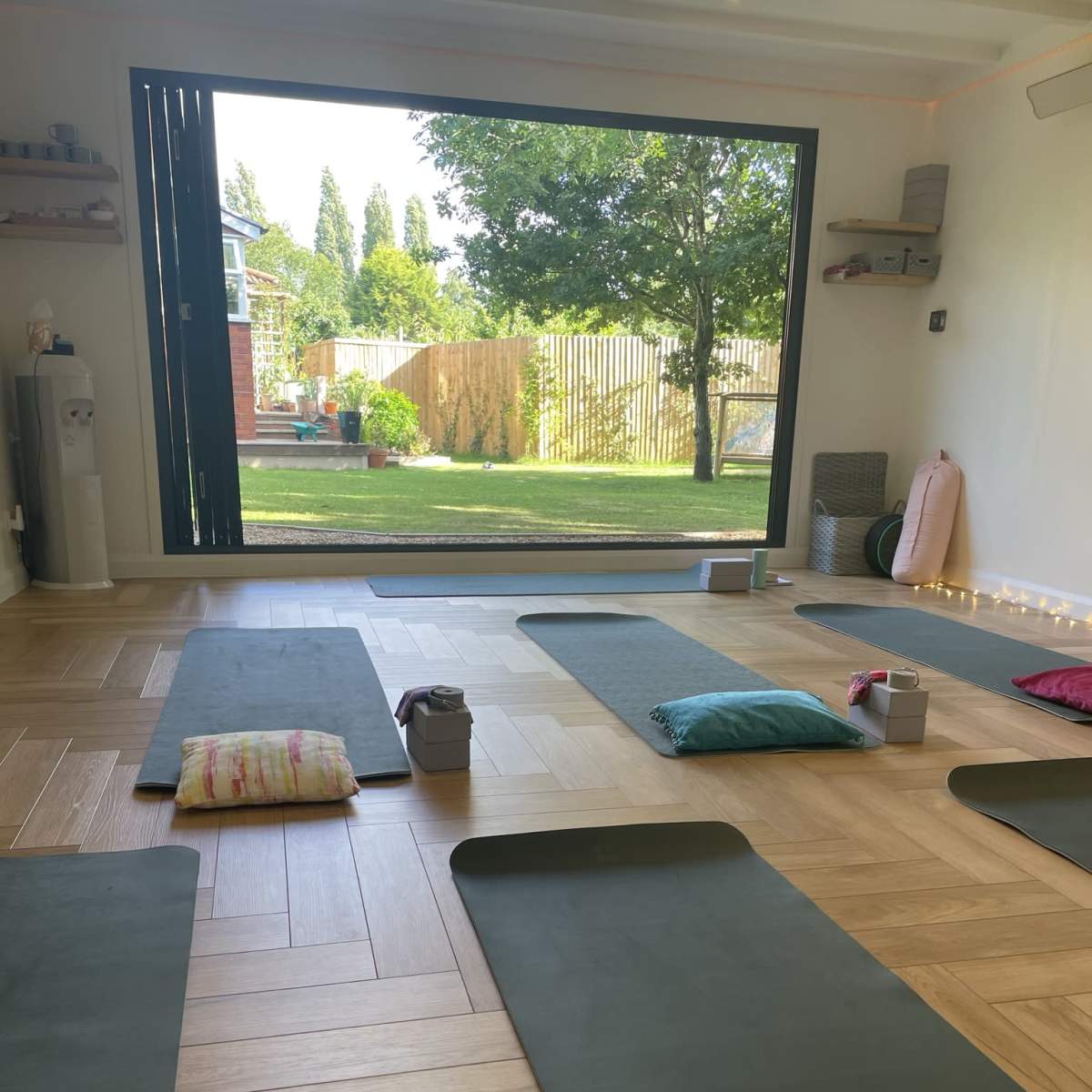 A yoga studio with green mats arranged on a wooden floor, overlooking a garden through a large window.