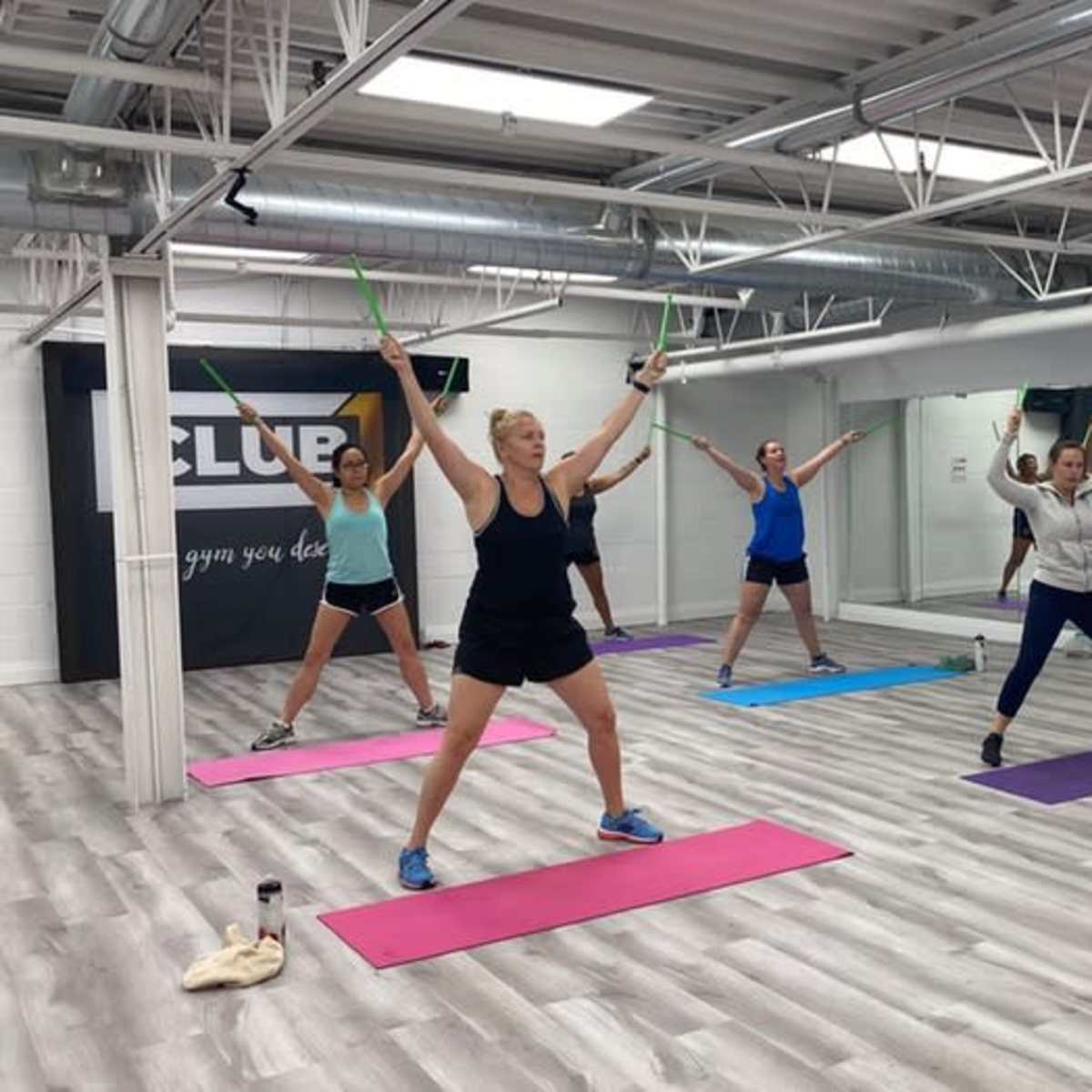 A group of individuals are participating in a fitness class, utilizing sticks while performing exercises on yoga mats in a mirrored studio.