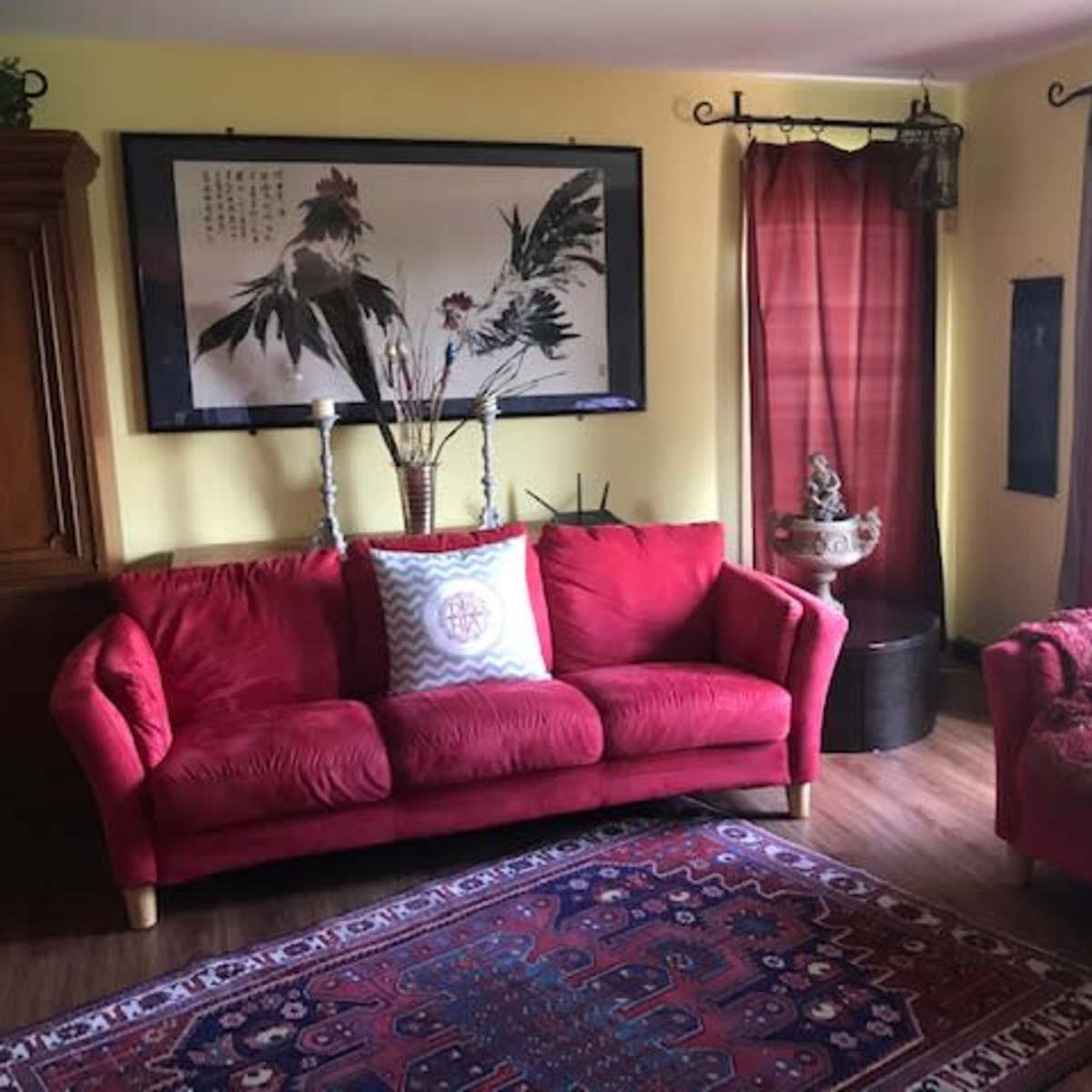 A living room with a red sofa, a small red armchair, a decorative rug, and a large framed artwork on the wall.