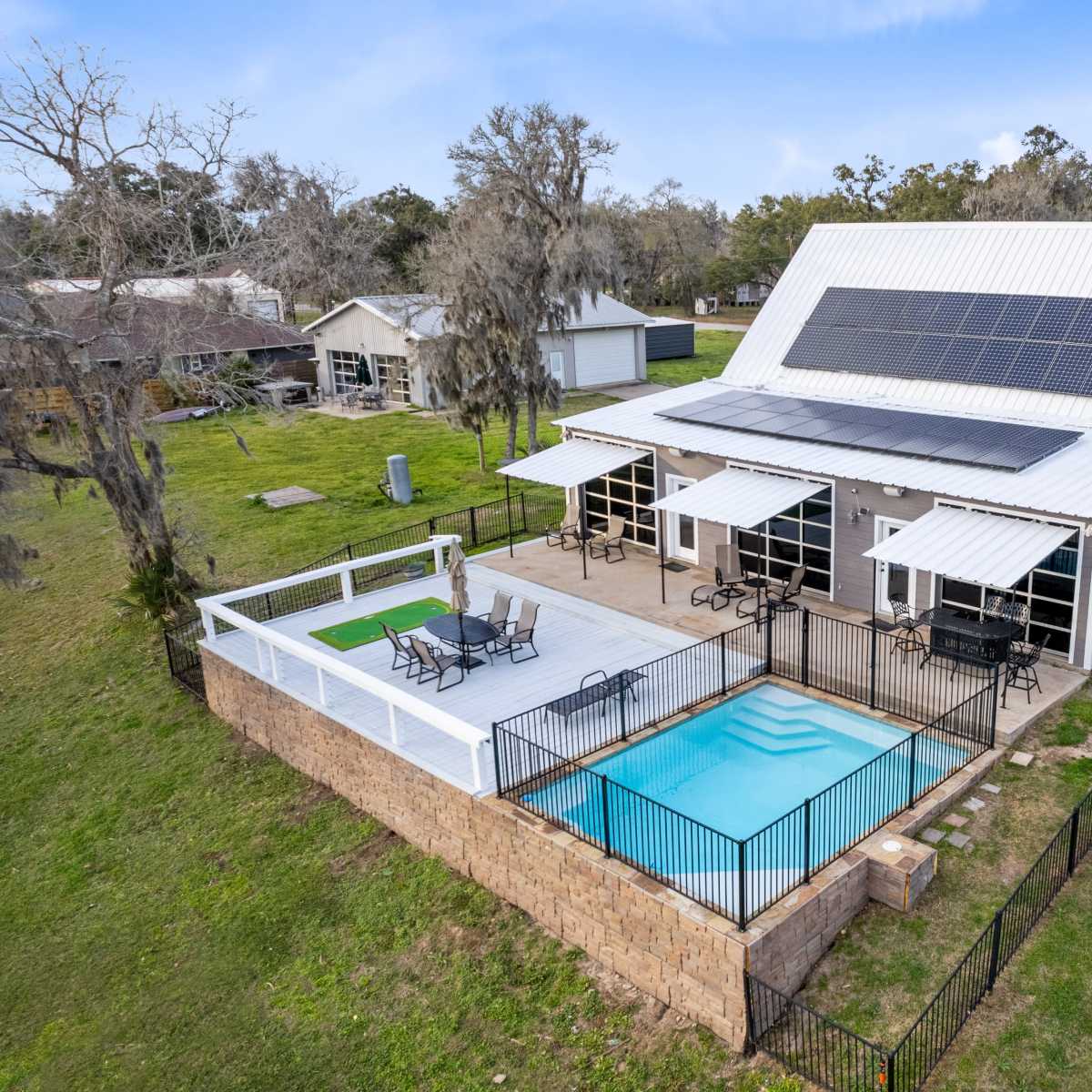 A modern house with a metal roof, solar panels, and a fenced swimming pool surrounded by a wooden deck and patio furniture.