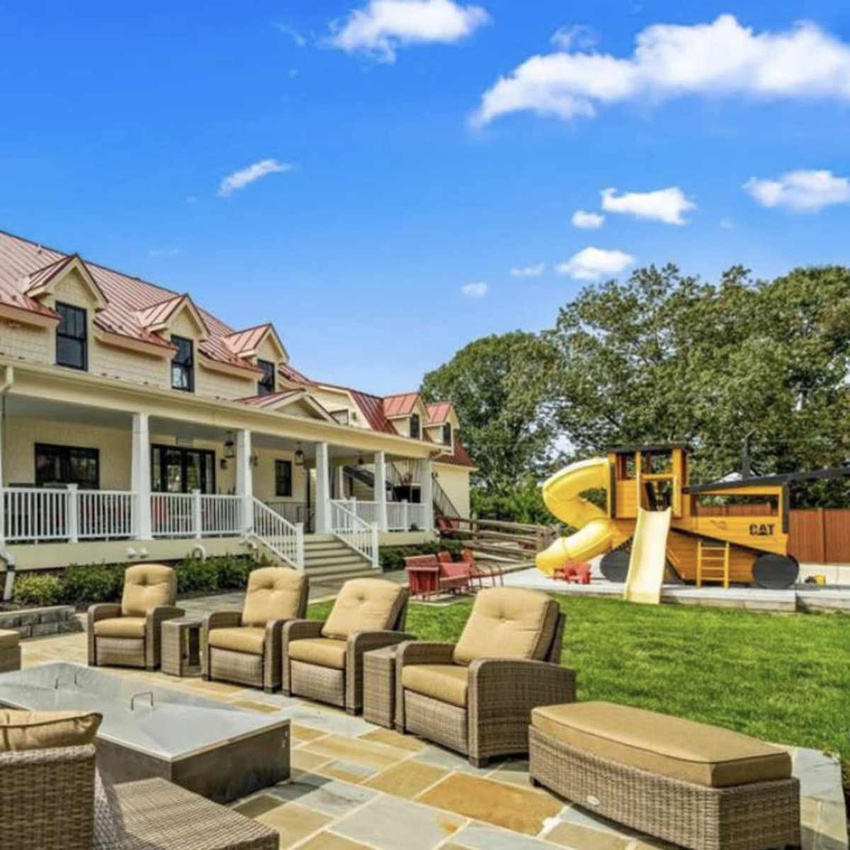 A spacious backyard features a seating area with wicker furniture surrounding a stone fire pit, alongside a large house with a red metal roof and a play structure.
