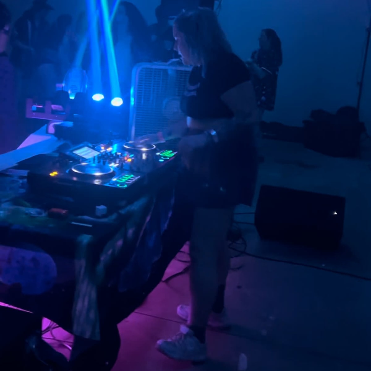 A DJ operates equipment with colorful lights while people dance in the background.