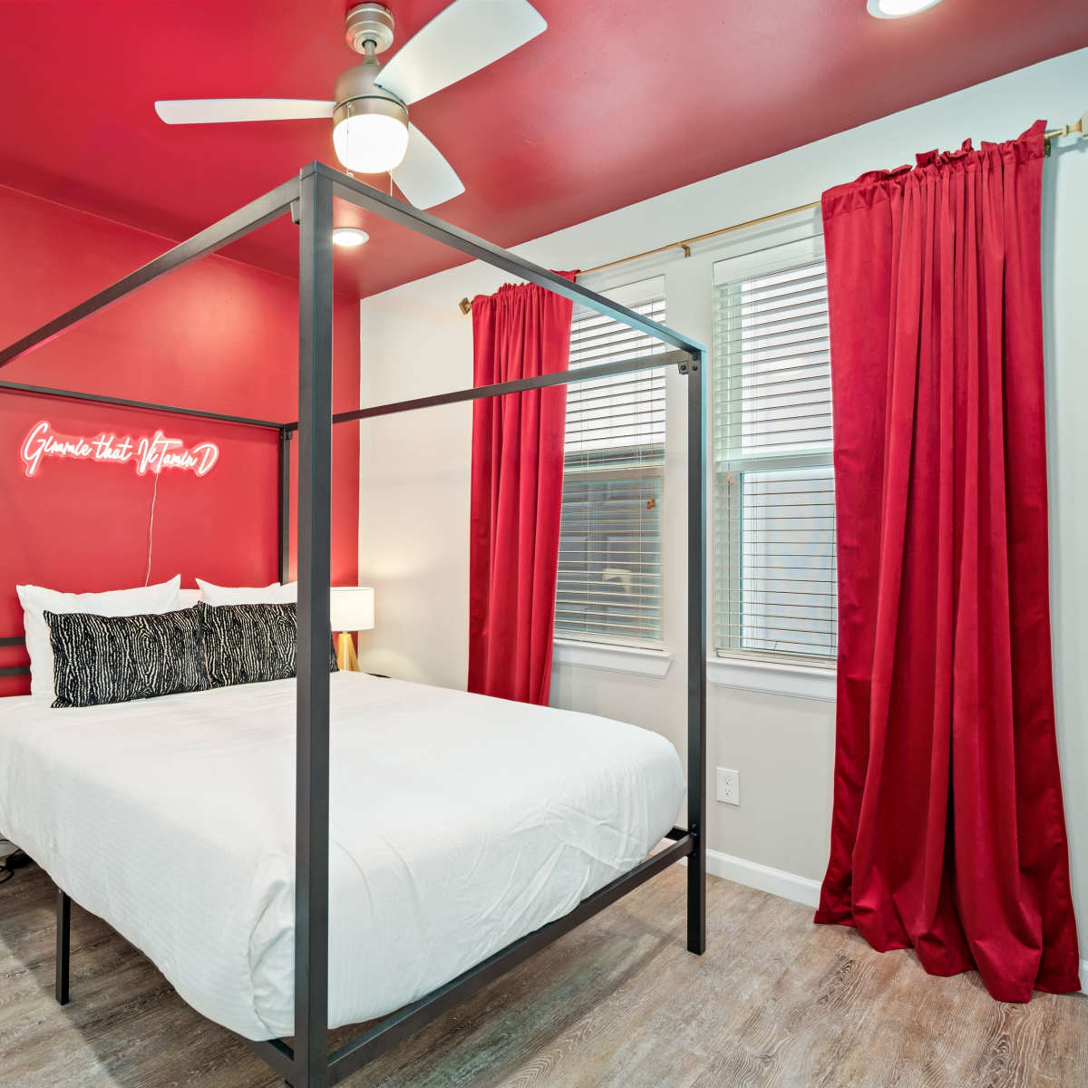 A bedroom features a four-poster bed with a white duvet, red walls, and large windows with red curtains.