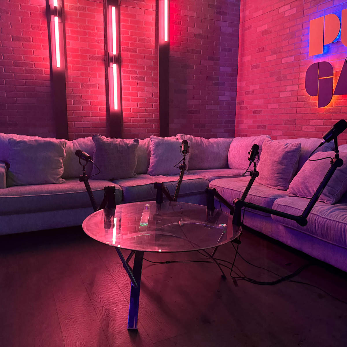 A modern lounge area features a gray couch and a glass coffee table, equipped with multiple microphones on stands, against a backdrop of illuminated brick walls.