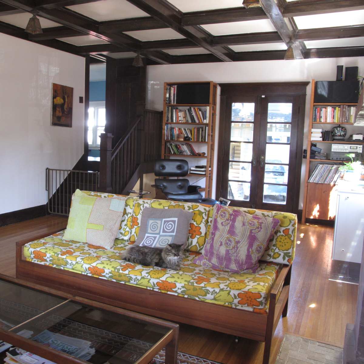 A cozy living room features a patterned couch, a coffee table, a bookshelf, and an open staircase leading to another level.