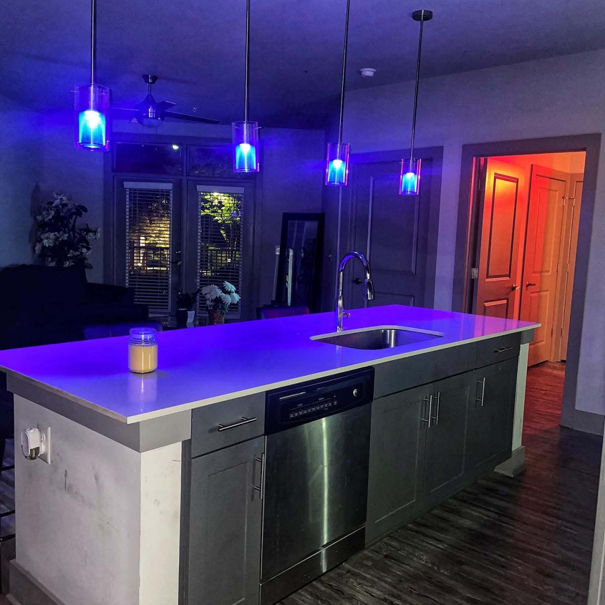 A modern kitchen features a central island with a sink, stainless steel dishwasher, and illuminated pendant lights casting blue and purple hues.