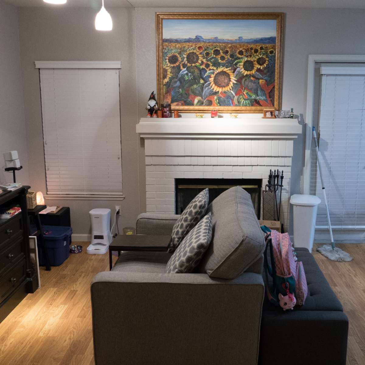 A cozy living room features a gray sofa, a television on a black cabinet, a fireplace adorned with a colorful sunflower painting, and a dining area with a table in the background.