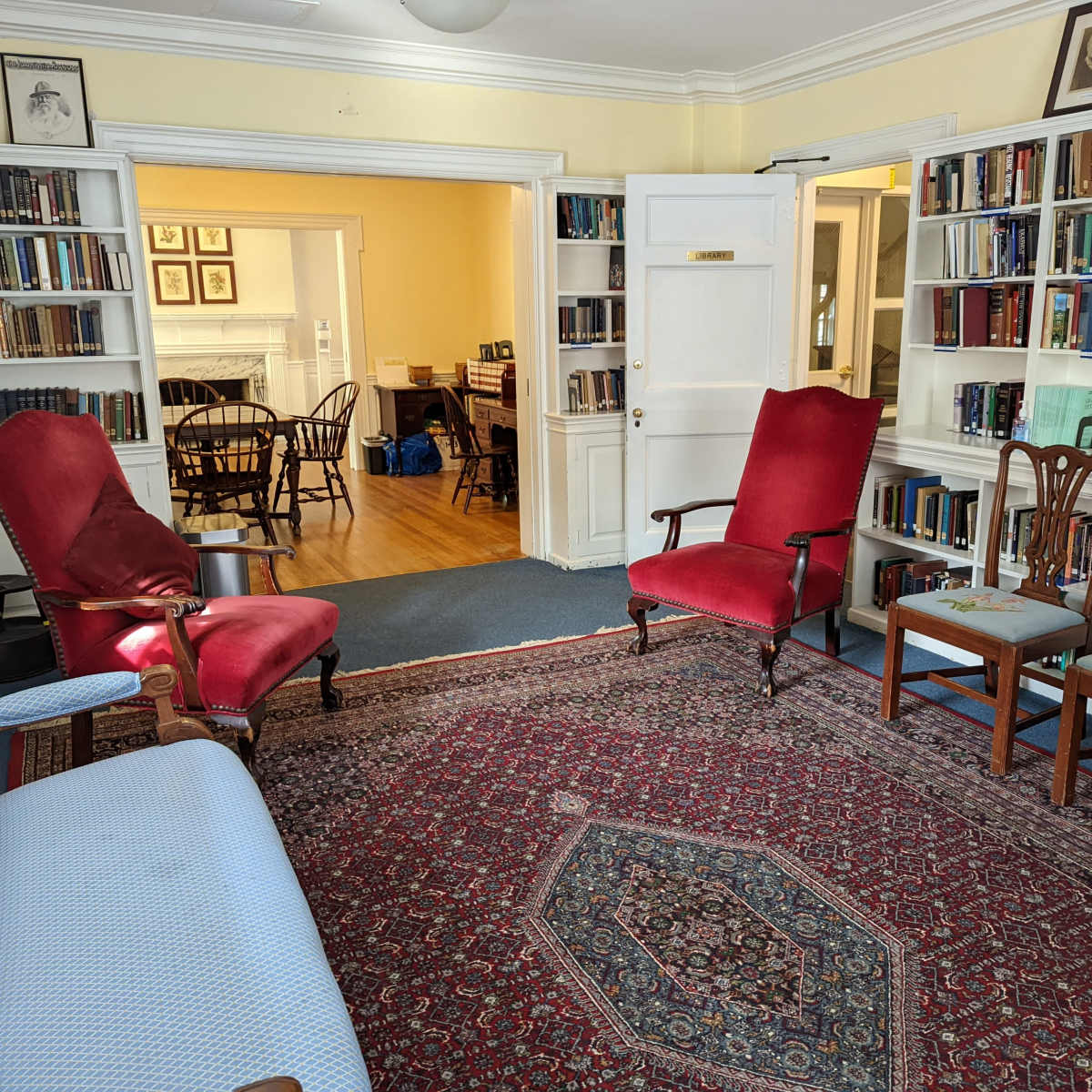A room with red upholstered armchairs, a patterned rug, and bookshelves filled with books, leading into another room with a dining area visible in the background.