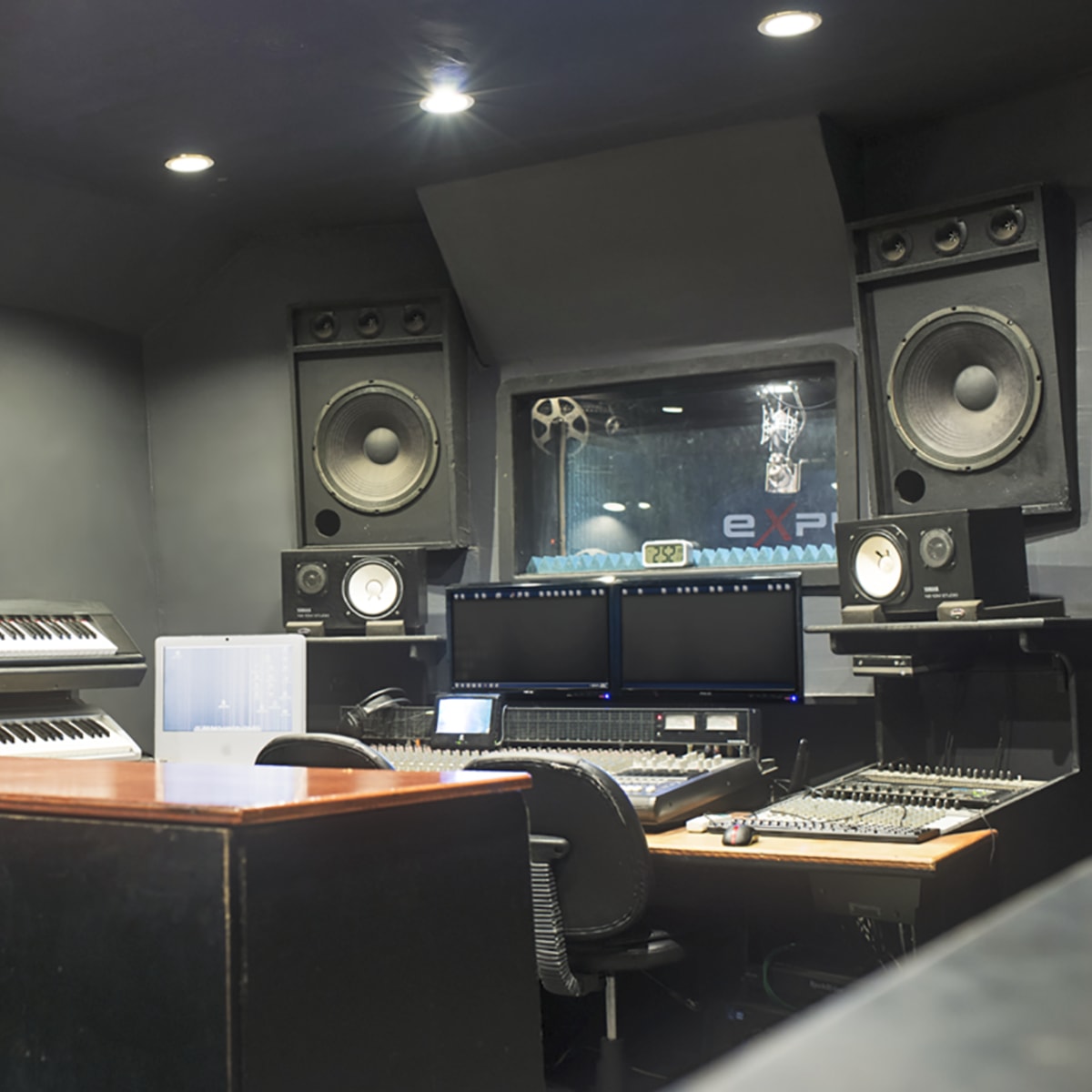 A music production studio featuring two computer monitors, a mixing console, synthesizers, and large studio speakers against dark walls.