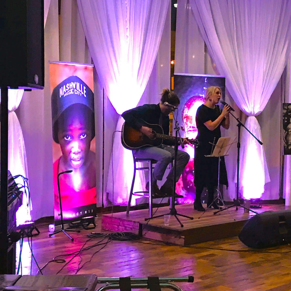 A woman sings into a microphone while a man plays guitar on a small stage, surrounded by ambient lighting and large banners featuring images.
