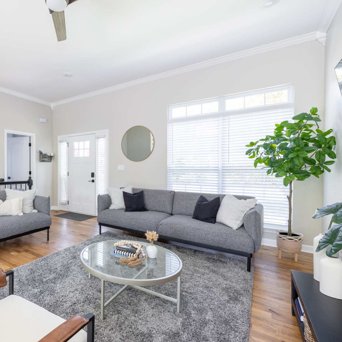 The living room features two gray sofas, a round glass coffee table, a rug, and a large window with white blinds.