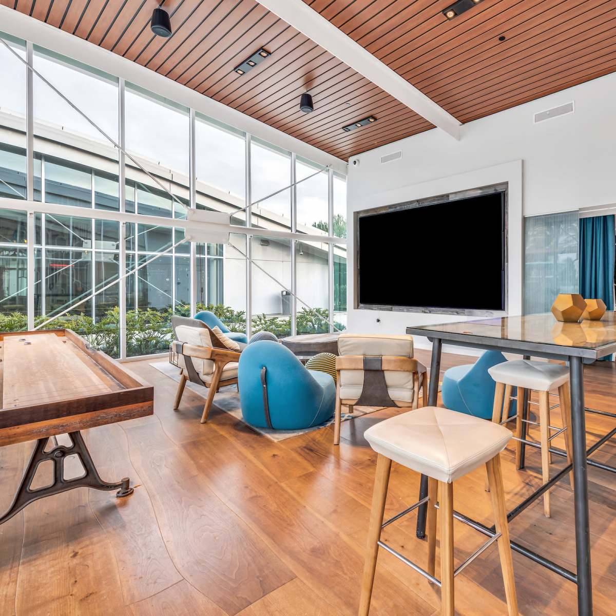A modern lounge area featuring a large screen, a wooden table, several stools, and bean bag chairs, with large windows allowing natural light.
