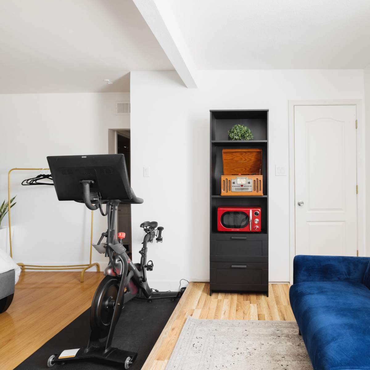 A small living space featuring a blue couch, a black shelving unit with a microwave, a stationary exercise bike, and a door leading to another room.