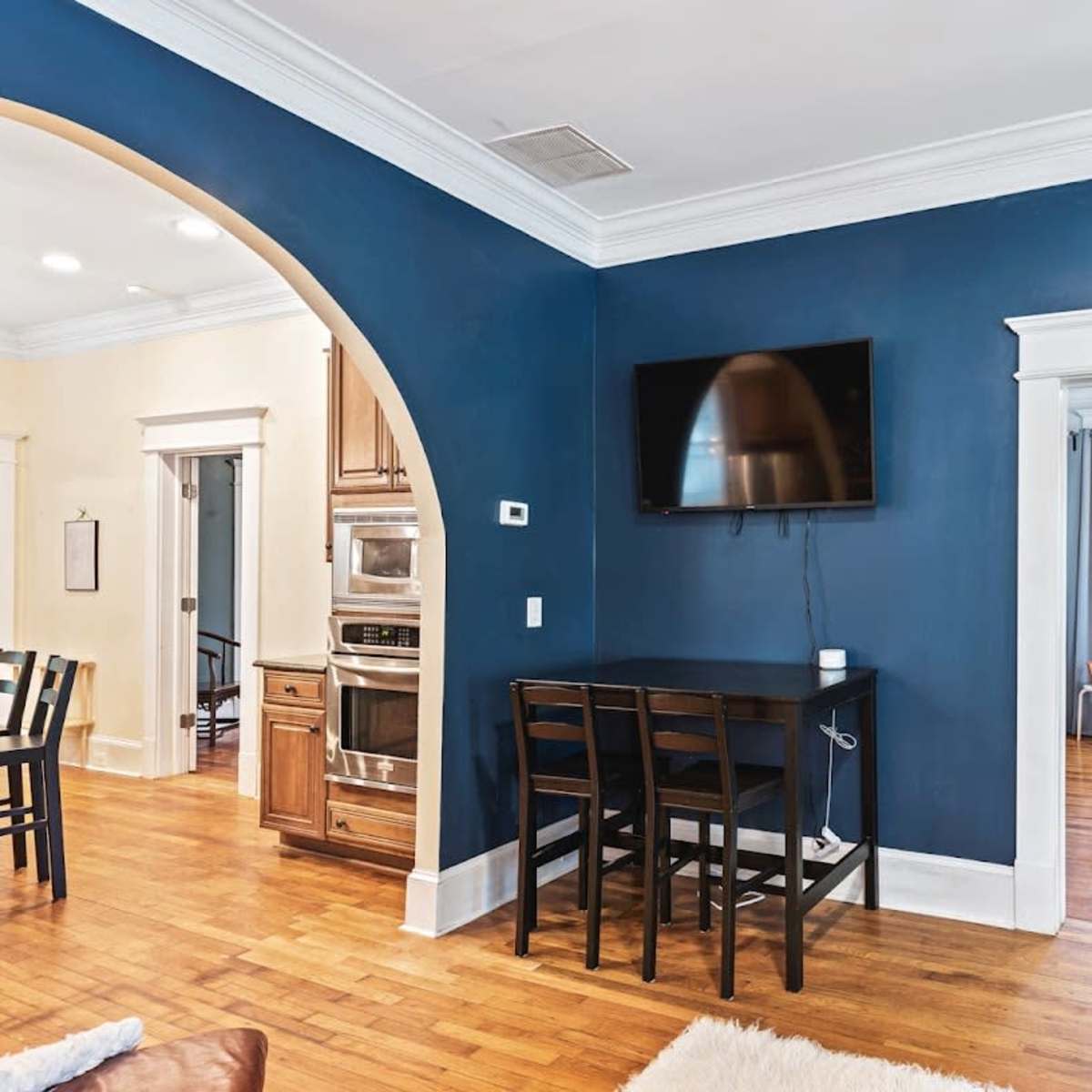 A living room with a dark blue wall, a mounted television, a small dining table with chairs, and a doorway leading to another room.