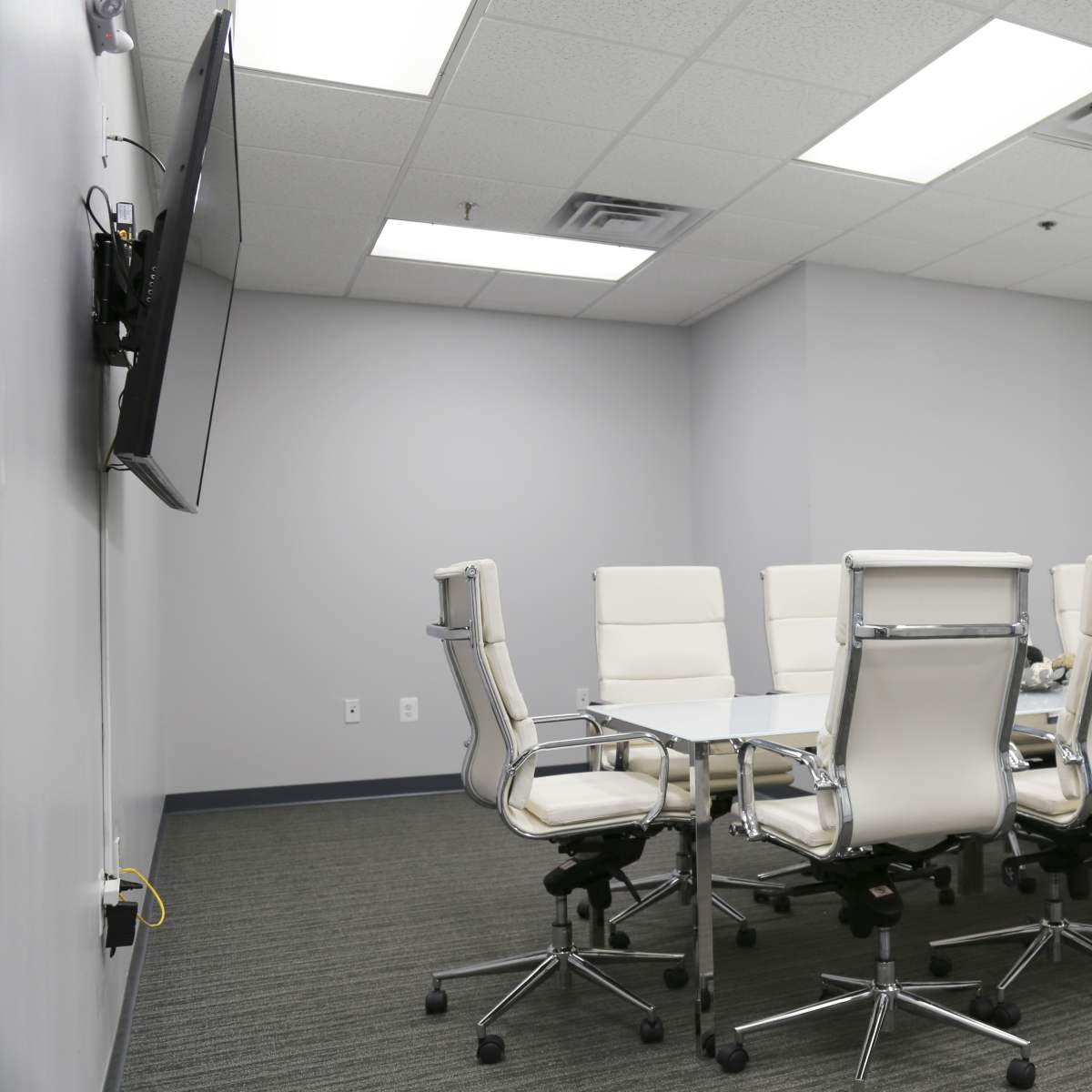 A modern conference room featuring a rectangular white table surrounded by eight white chairs and a wall-mounted television.